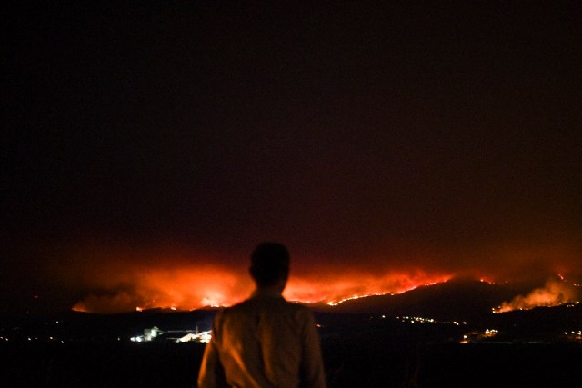 A man stands on the roadside and watches a wildfire at Anciao, Leiria, central Portugal, on June 18, 2017. A wildfire in central Portugal killed at least 25 people and injured 16 others, most of them burning to death in their cars, the government said on June 18, 2017. Several hundred firefighters and 160 vehicles were dispatched late on June 17 to tackle the blaze, which broke out in the afternoon in the municipality of Pedrogao Grande before spreading fast across several fronts. / AFP PHOTO / Patricia De Melo MOREIRA