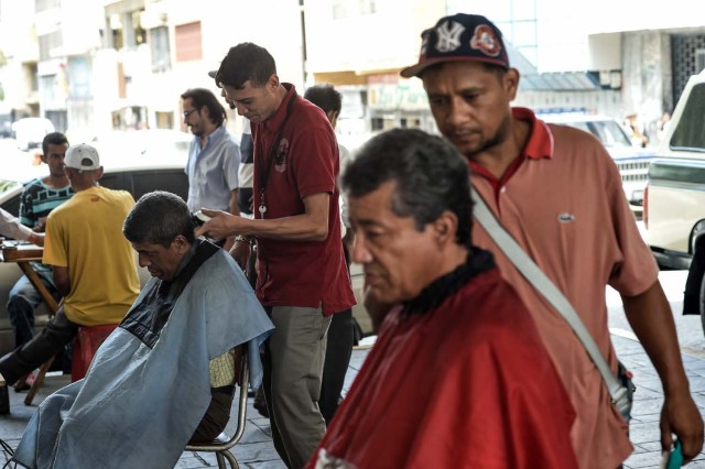 Dos hombres trabajan cortando pelo bajo un puente en Caracas el 28 de mayo de 2018. Bajo un puente o en un mercado al aire libre en Caracas, los peluqueros se ganan la vida con herramientas simples y ofrecen cortes de pelo para aquellos que no pueden pagar un salón de belleza porque de la crisis económica. / AFP PHOTO / Juan BARRETO