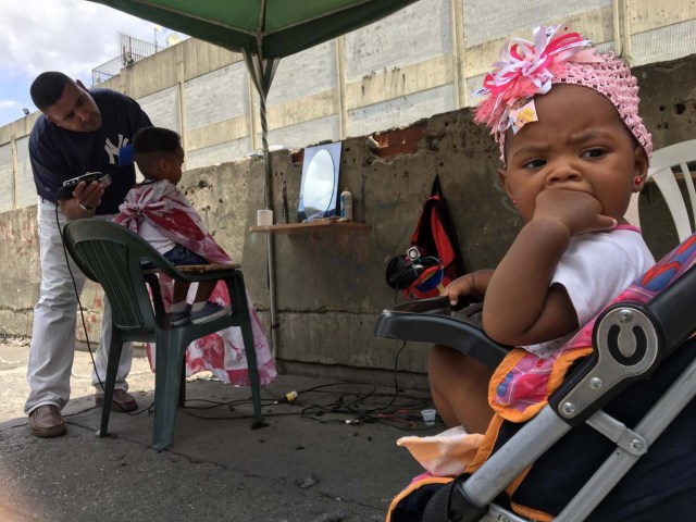 Un hombre corta el cabello de un niño en una calle de Caracas el 4 de junio de 2018. Bajo un puente o en un mercado al aire libre en Caracas, los peluqueros se ganan la vida con herramientas simples y ofrecen cortes de pelo para aquellos que no pueden pagar un salón de belleza salón de belleza debido a la crisis económica. / AFP PHOTO / Juan Barreto